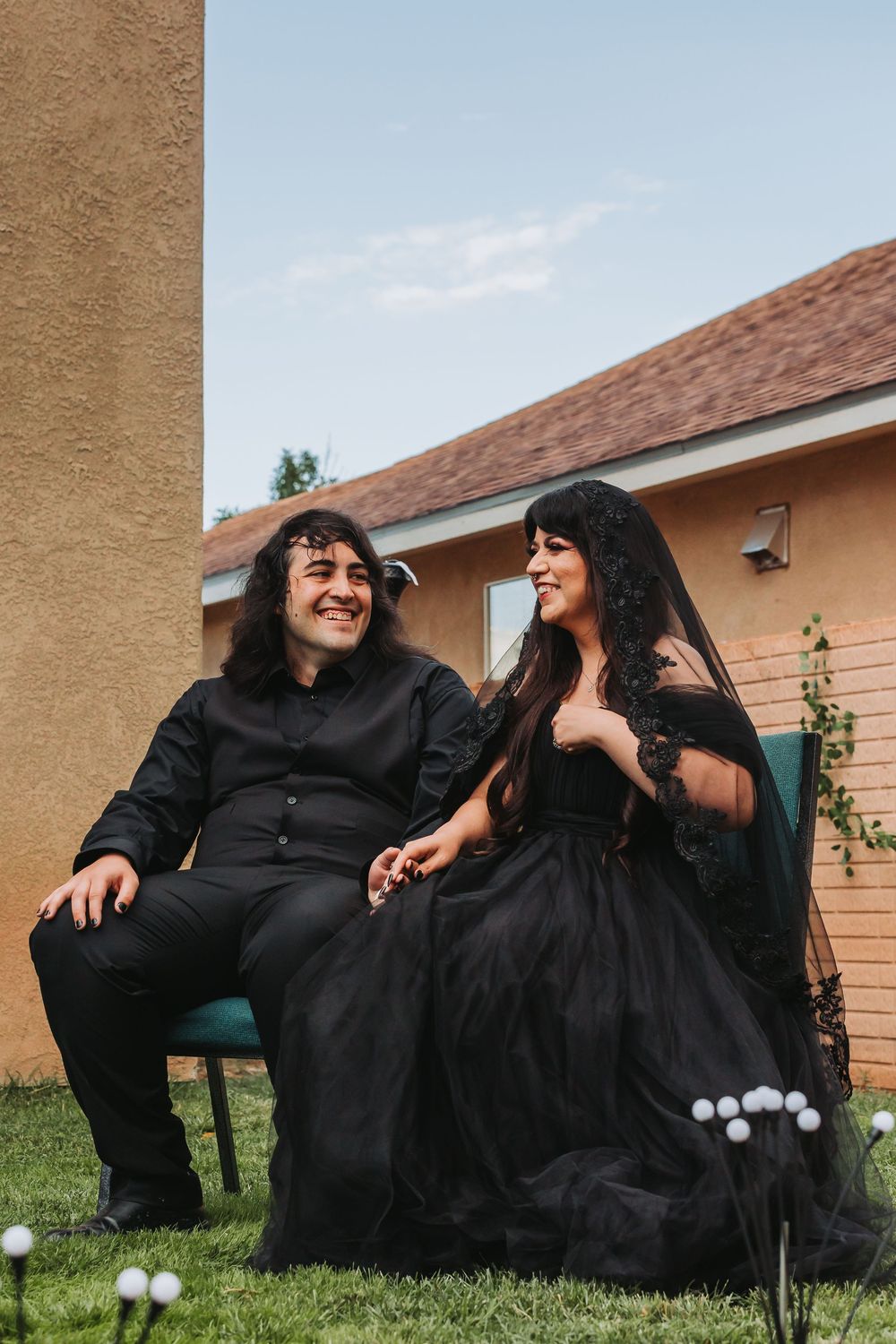 Two people wearing black robes laugh together while sitting on grass outside a house.