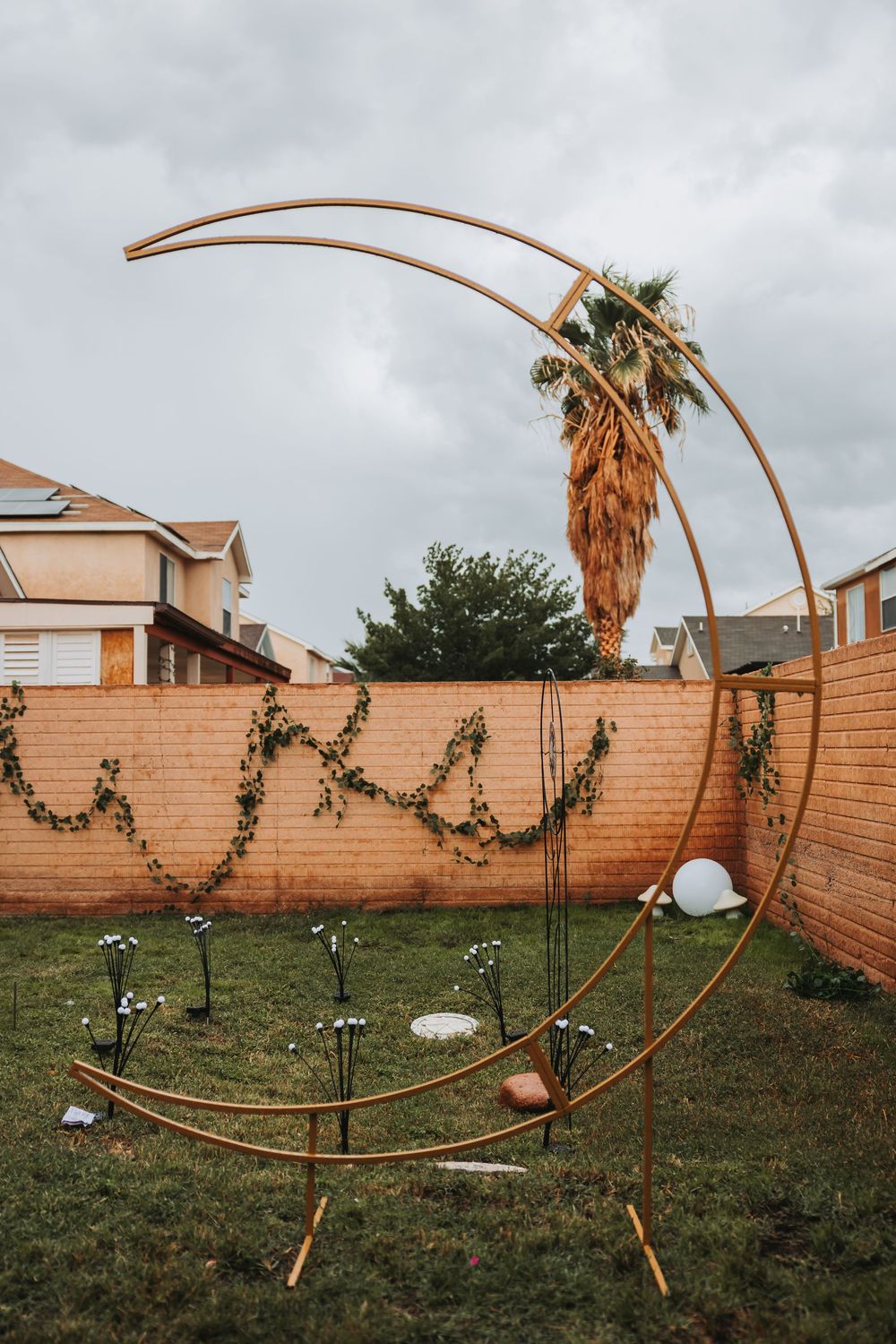 A curved wooden arch installation with hanging greenery stands in a backyard against a brick wall and palm trees.