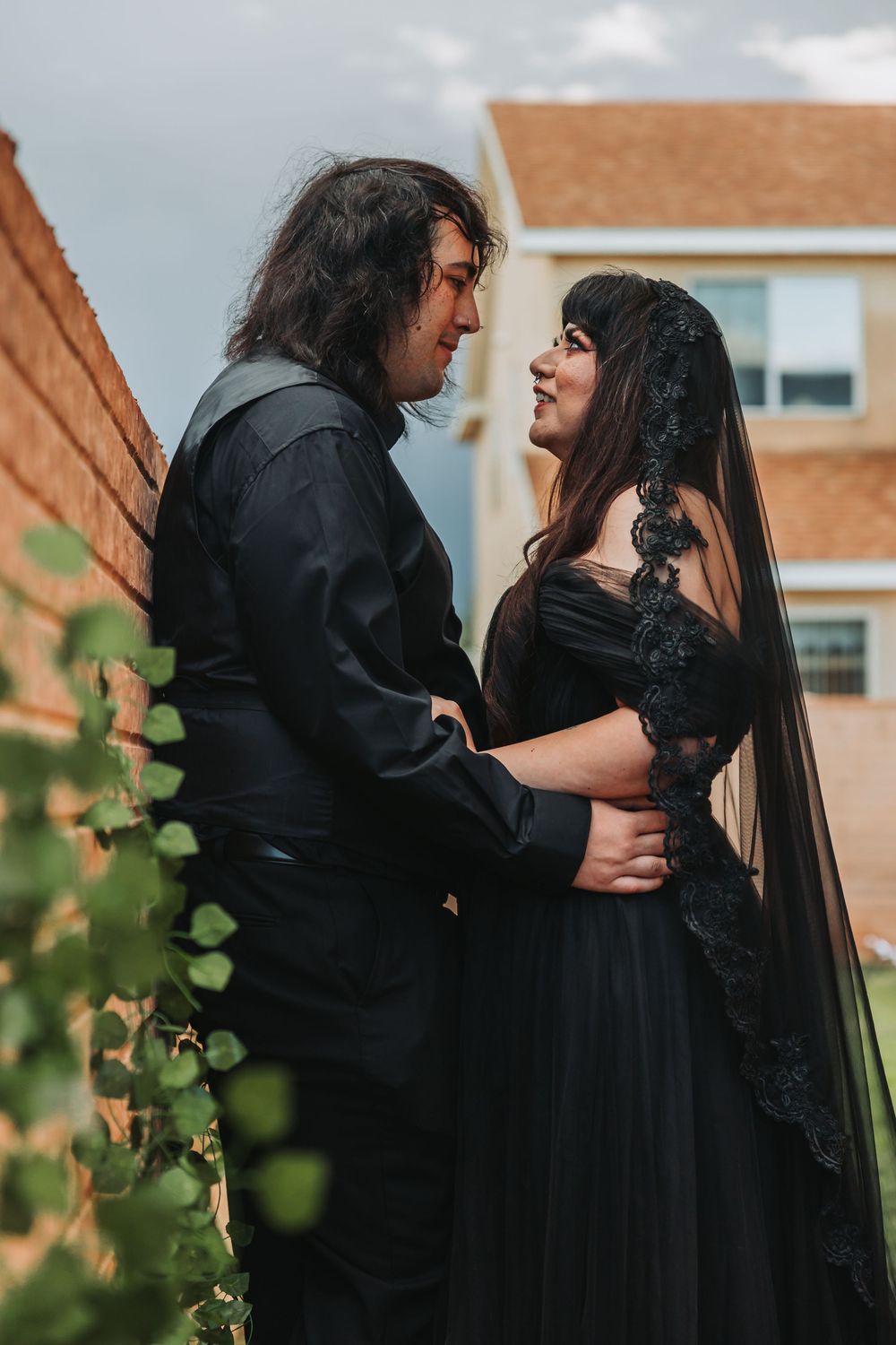 A couple in black outfits embrace in a romantic outdoor photoshoot series with brick buildings in the background.