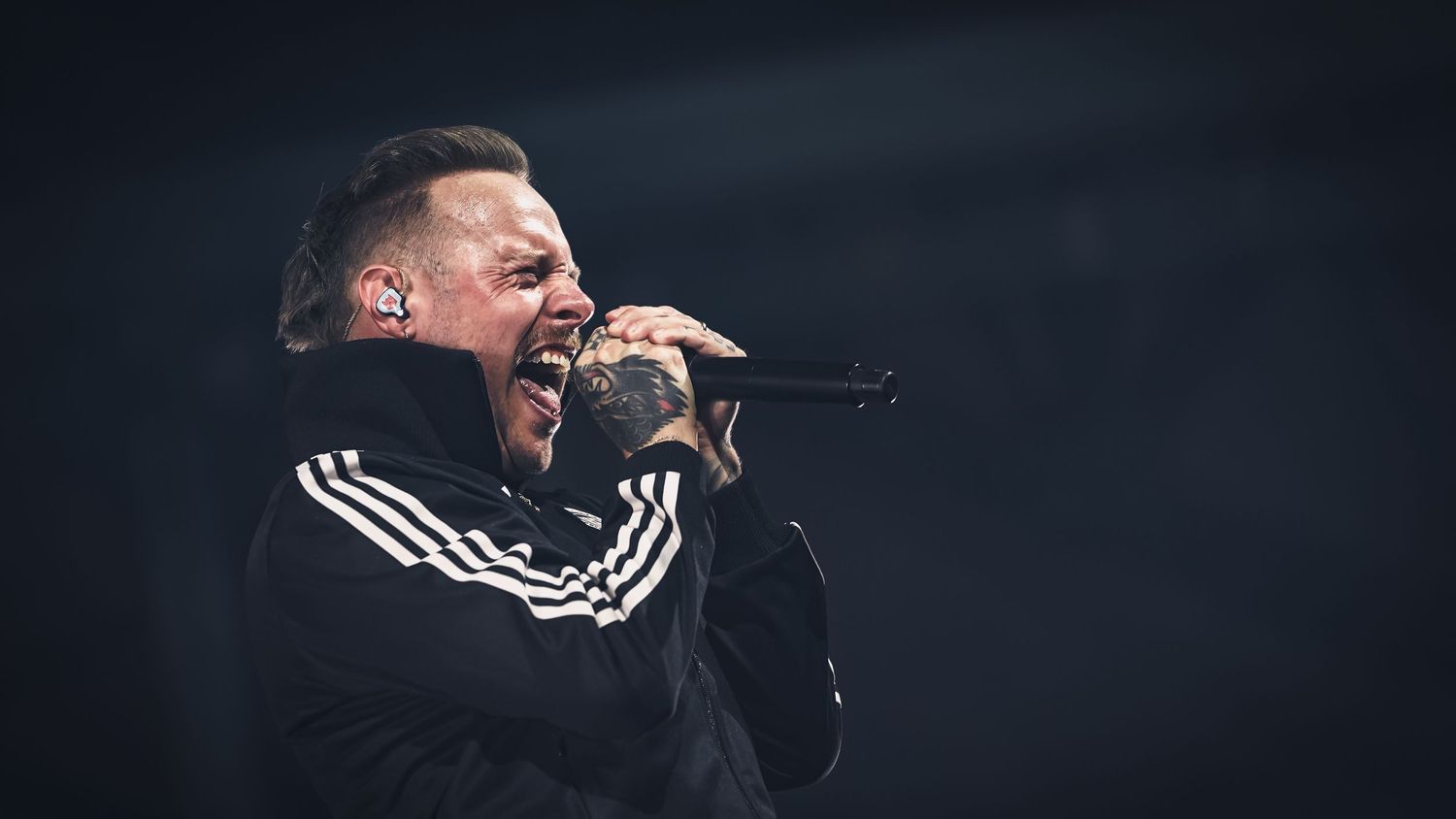 Performer in black clothing singing passionately on a dark stage with dramatic lighting.