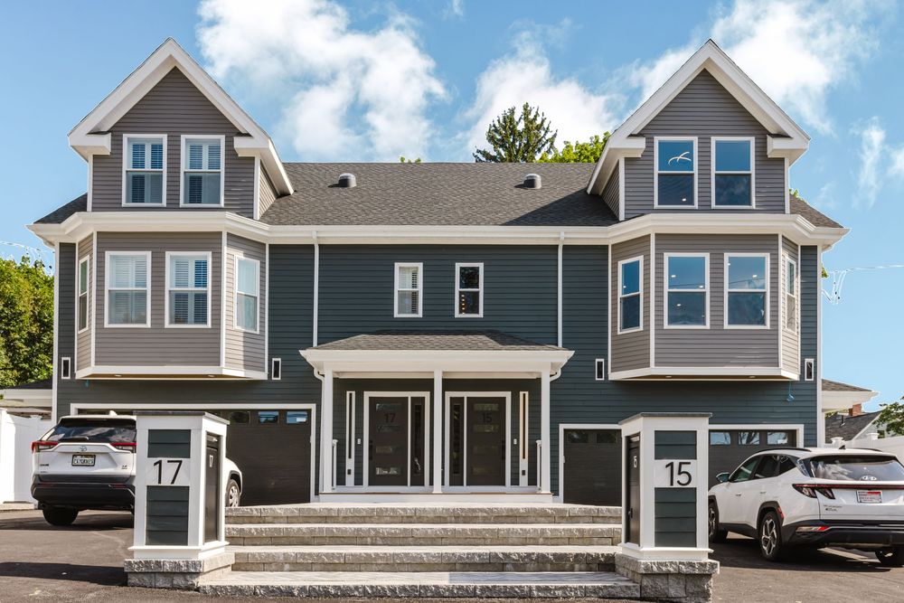 Modern gray two-story colonial home with bay windows, white trim, covered entry stairs, and numbers 15 and 17 visible.