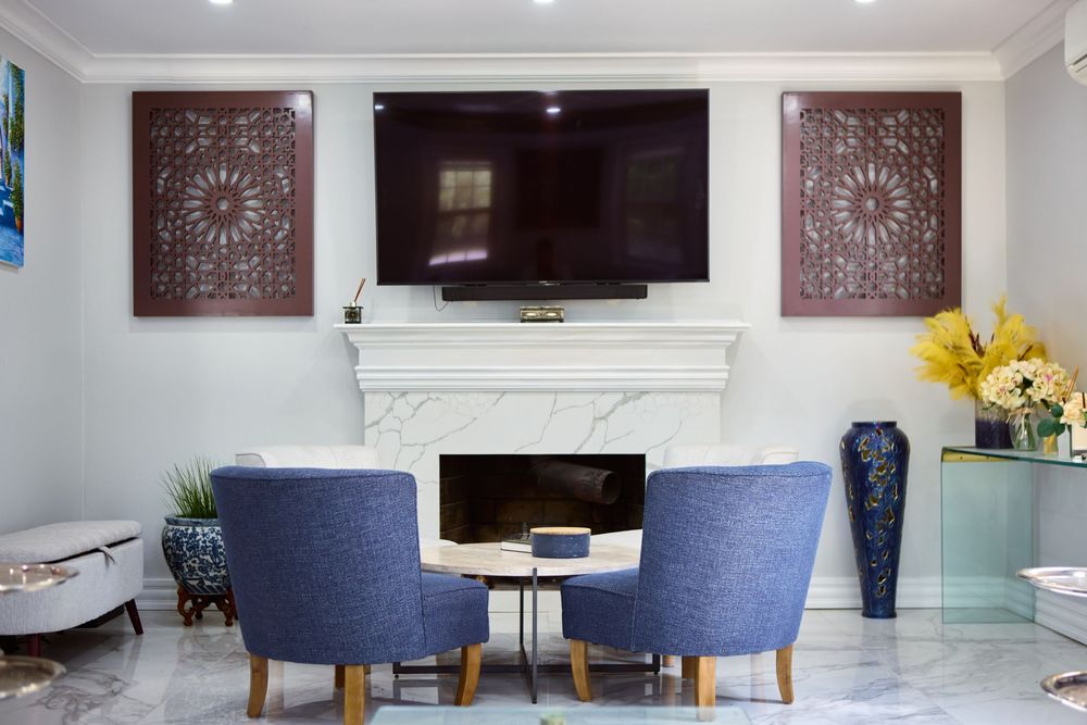 Elegant living room with white marble fireplace, mounted TV, blue accent chairs, and decorative carved wood wall panels.