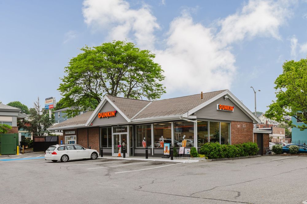 Dunkin Donuts corner retail location with brick exterior, large windows, parking lot, and mature trees on a sunny day.