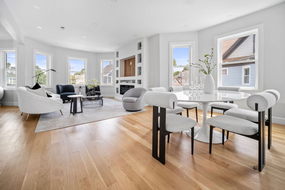 Open-concept living and dining area with hardwood floors, bay windows, and modern white furnishings.