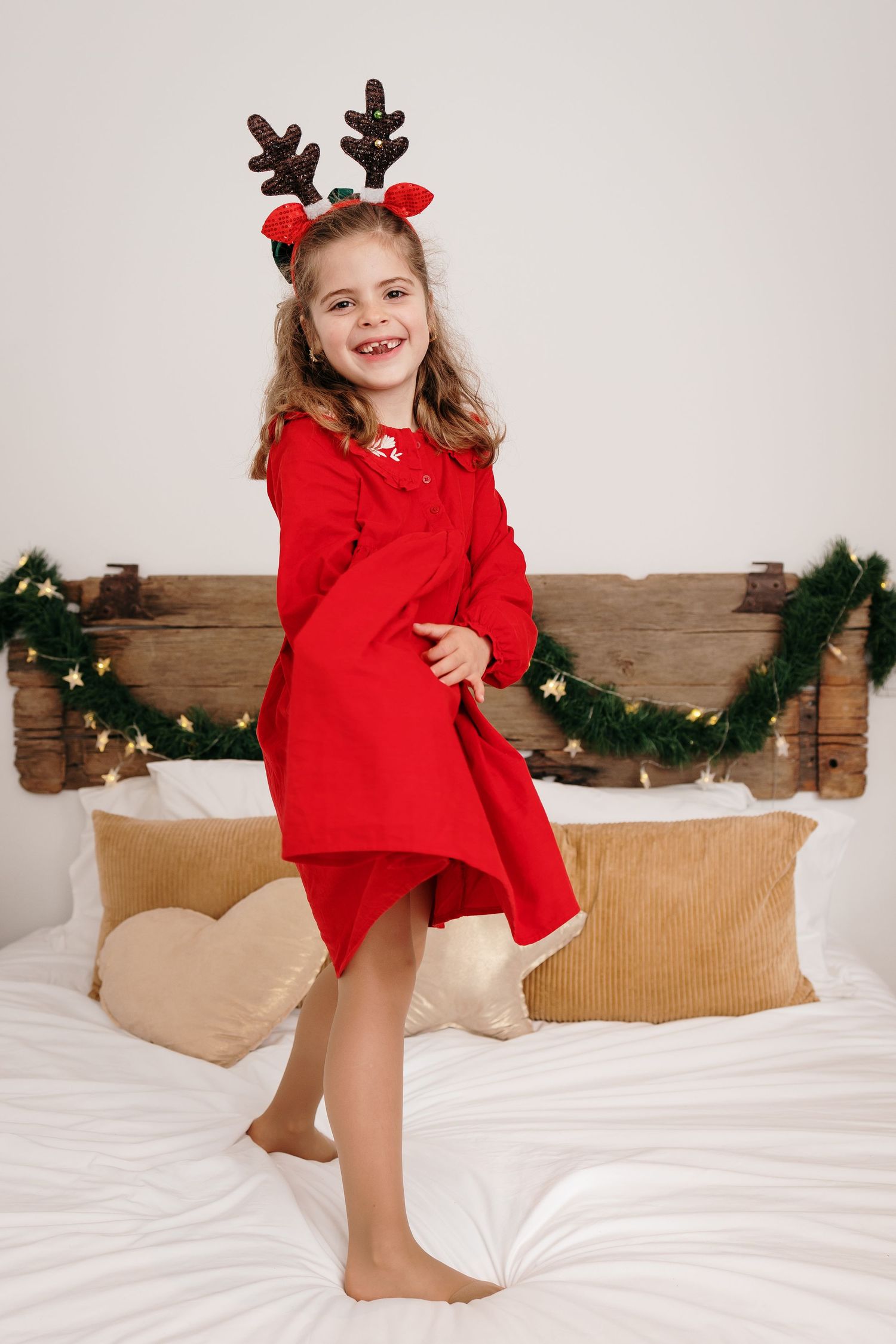 Child in red holiday outfit jumping playfully on white bedding with festive garland decor.