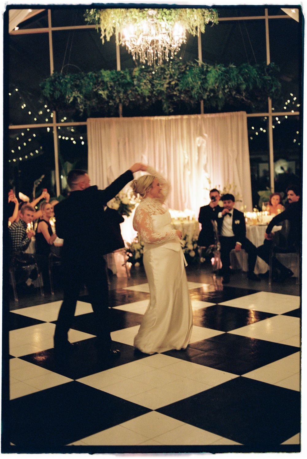 A couple dances elegantly on a black and white checkered dance floor under crystal chandeliers at an evening reception.