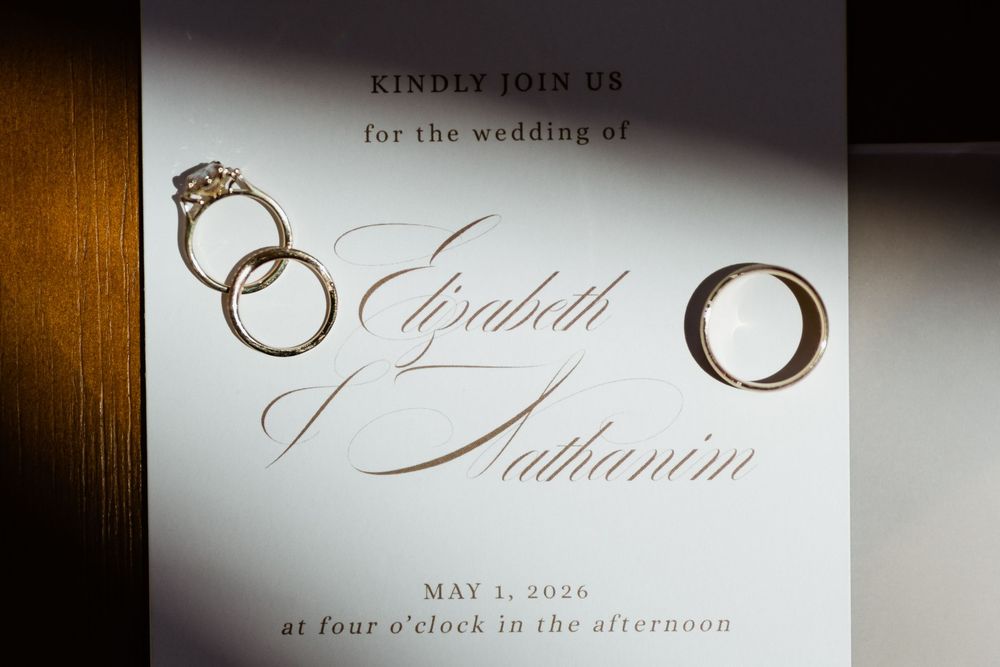 Elegant wedding invitation for Elizabeth and Nathanim on May 1, 2026, displayed with gold wedding rings on white background.