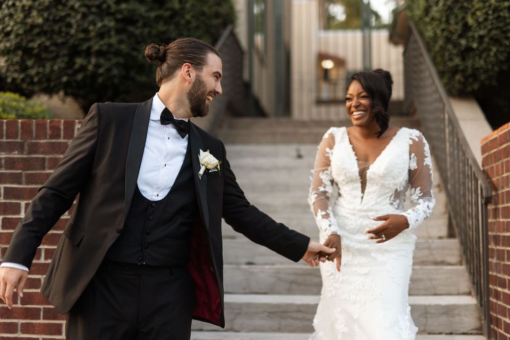 Newlywed couple shares joyful moments during outdoor wedding photoshoot against brick architecture.