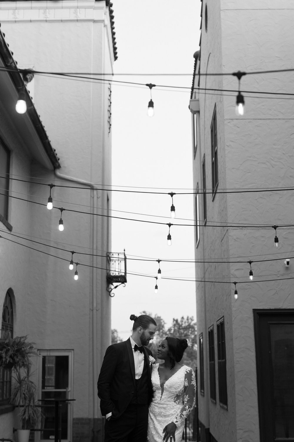 String lights illuminate a modern outdoor courtyard space between white painted buildings at dusk.
