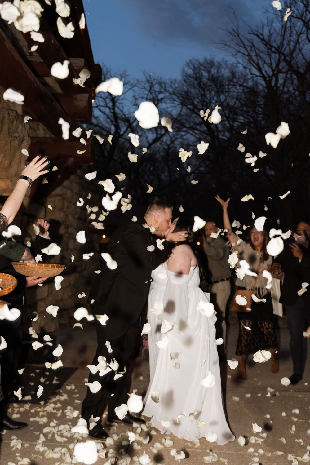 A couple embraces during their wedding celebration as white rose petals fall around them in an outdoor evening setting.