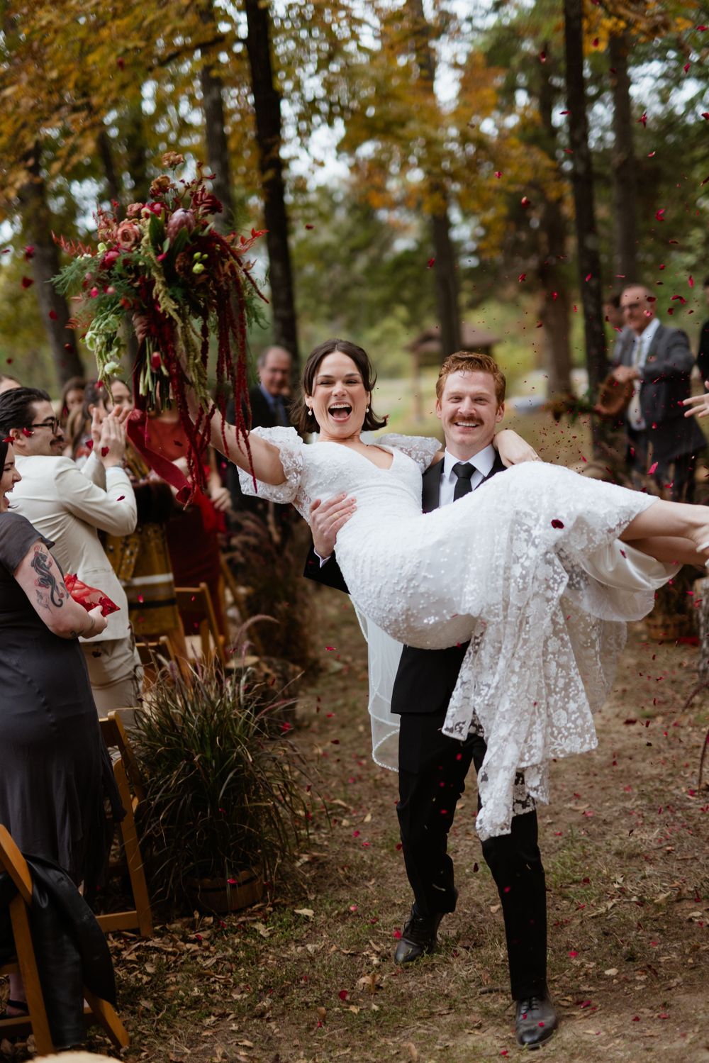 A groom carries his bride in a white lace dress through a wooded outdoor wedding ceremony setting during autumn.