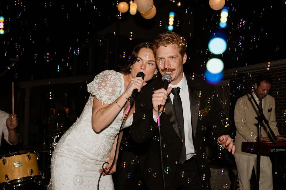 Bride and groom sing karaoke together at their wedding reception while holding microphones under glowing lights.
