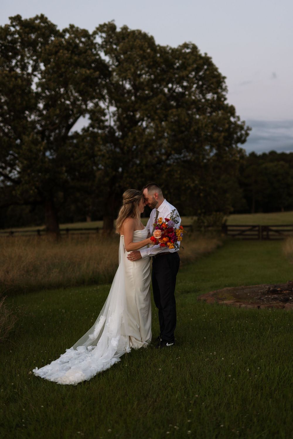 A romantic wedding couple embraces at sunset in a field with large oak trees in the background.