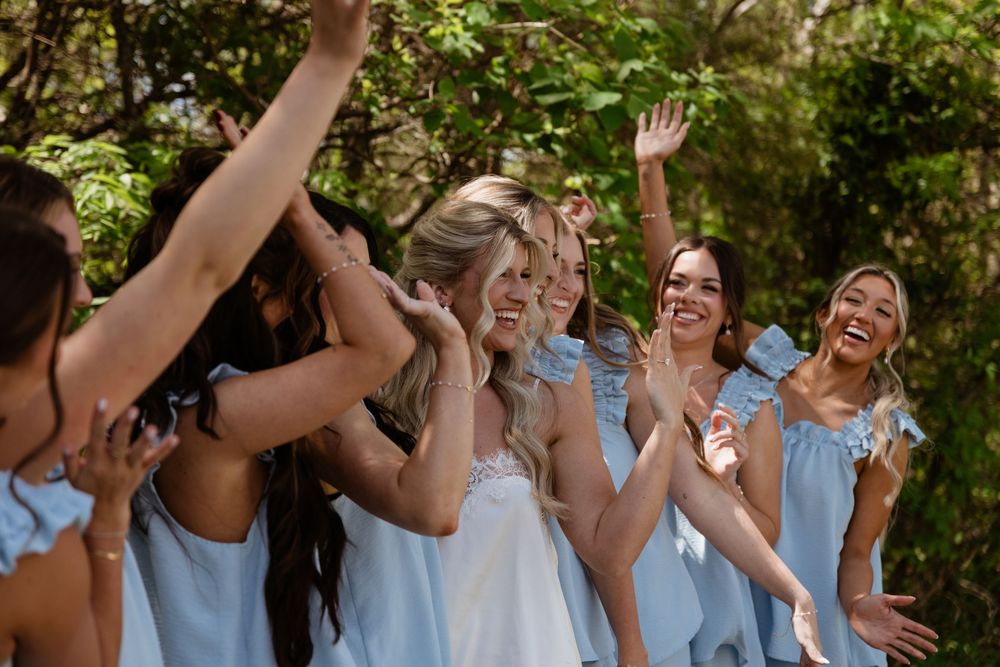 Bride and bridesmaids in light blue dresses laughing and celebrating outdoors surrounded by lush green trees.