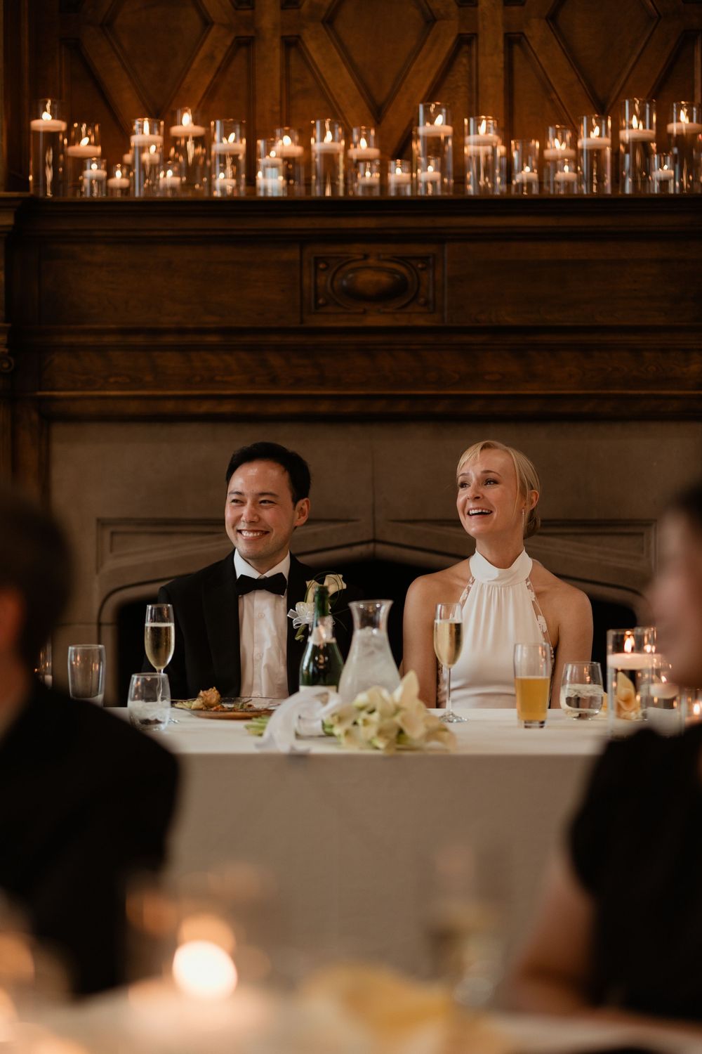 Wedding reception guests share laughter and conversation at an elegant candlelit dinner table in a grand ballroom.