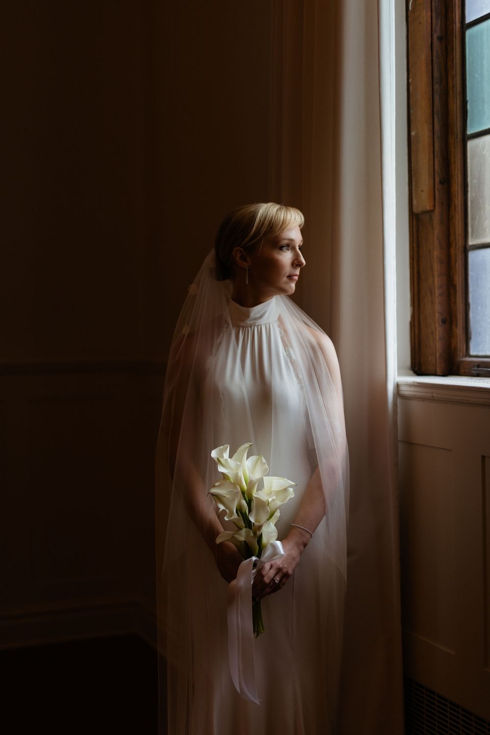 Bride in flowing white veil holds elegant white lily bouquet while standing near window in dramatic lighting.