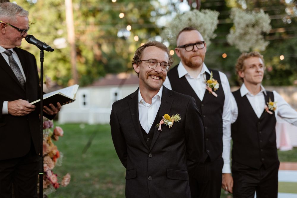 A group of groomsmen in black suits and ties share a candid moment outdoors during a wedding celebration.