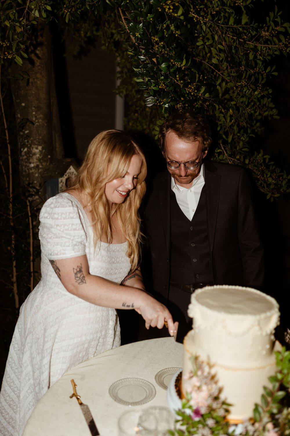 A couple cuts into their white wedding cake together during an evening outdoor reception celebration.