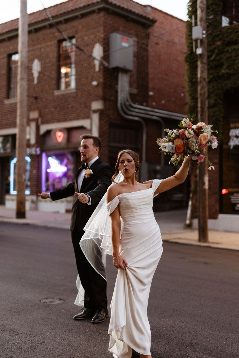 A bride and groom dance playfully on an urban street at dusk with brick buildings in the background.