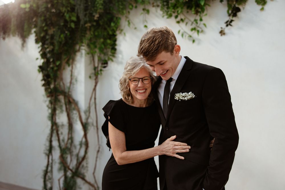 A romantic couple embraces against a white wall with climbing vines in formal black attire.