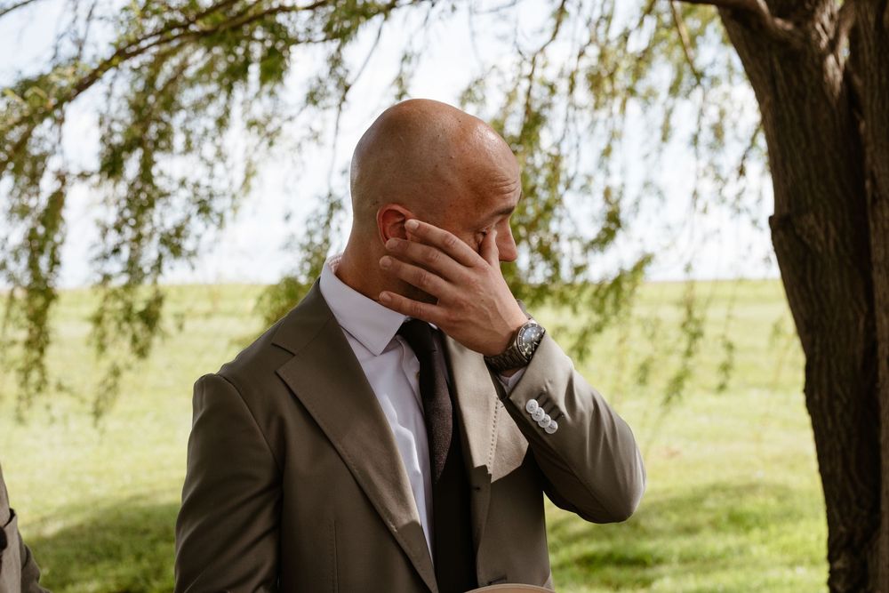 A person in a brown suit covers their face with their hand while standing outdoors under a willow tree.