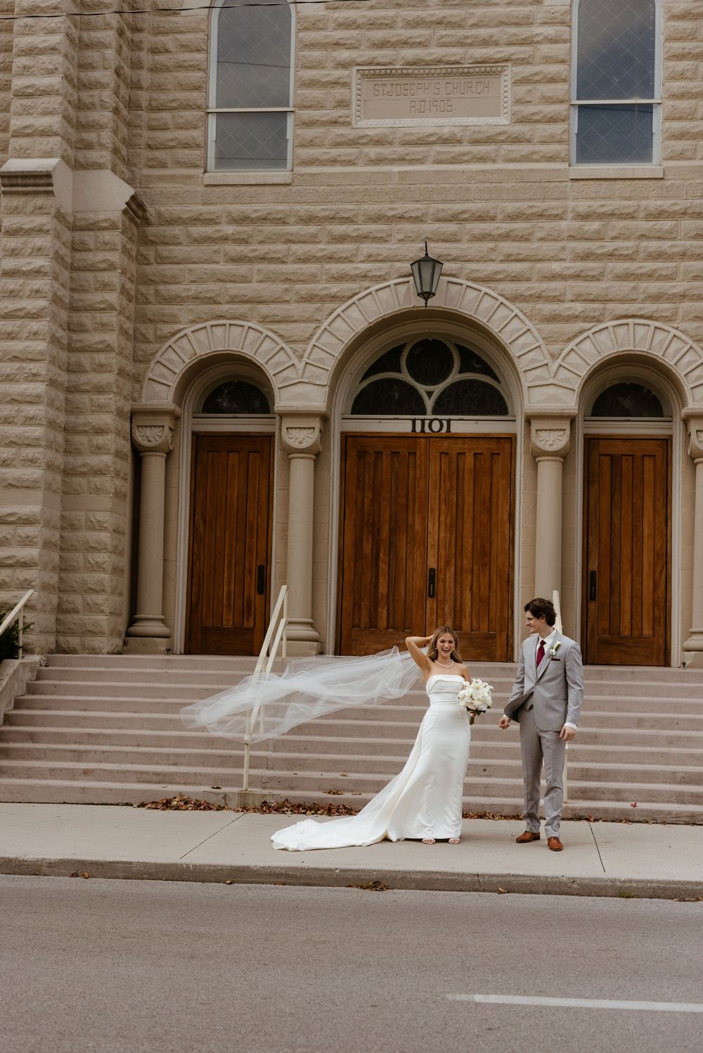 A wedding couple poses outside a stone building as the bride's veil flows dramatically in the wind.