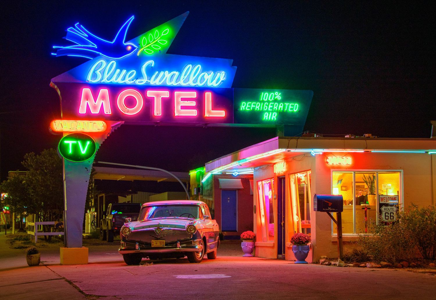 Vintage neon motel signs illuminate the night sky with vibrant blue, green and red colors along a roadside strip.