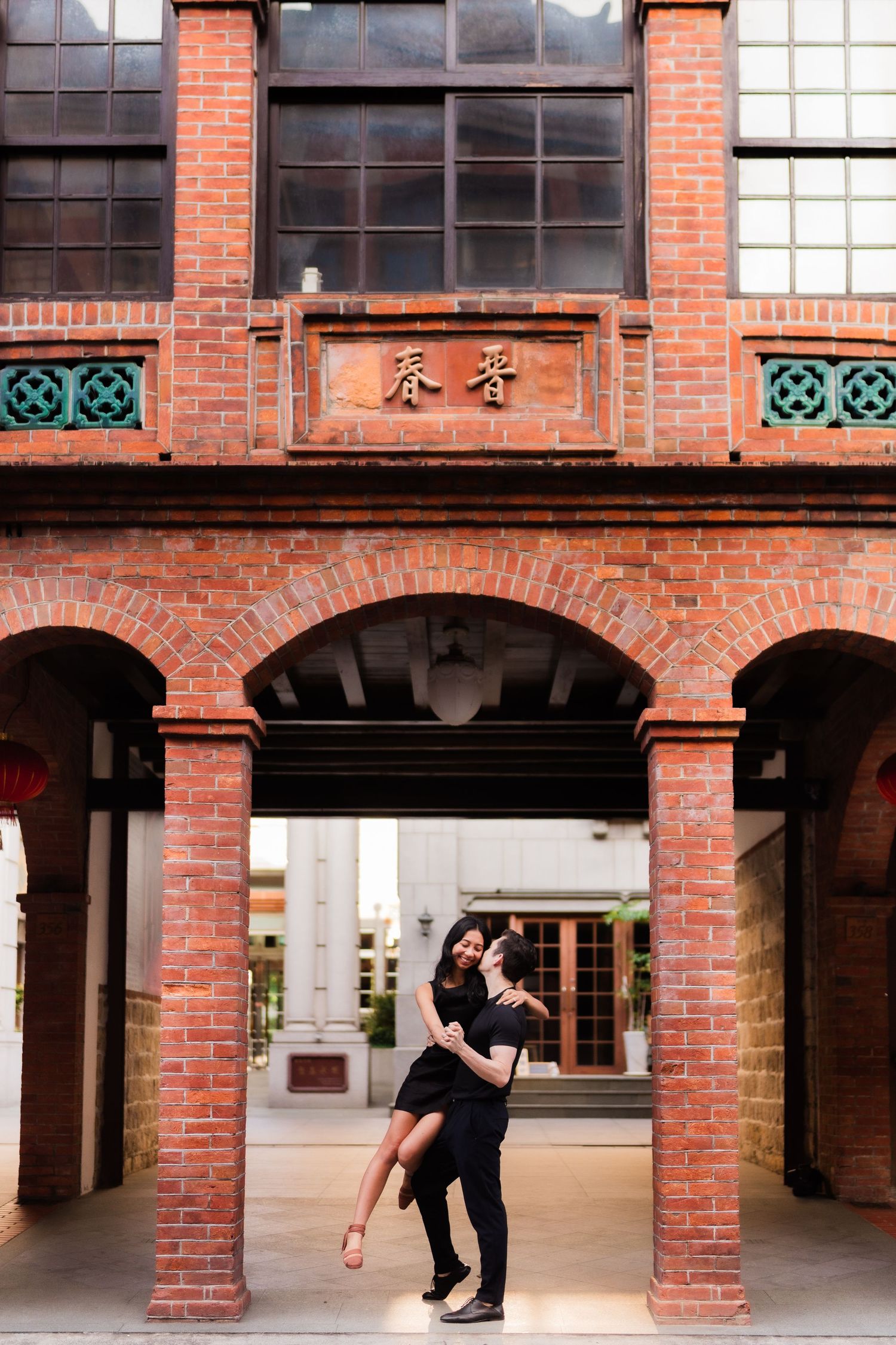 A couple dances under a red brick archway with Chinese characters and decorative teal accents at a historic building.