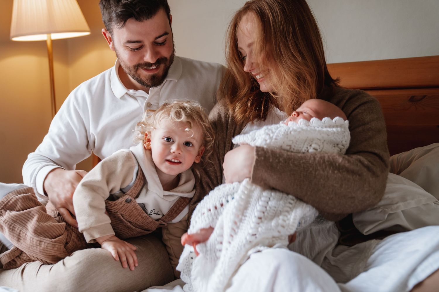 Happy family of four with toddler and newborn baby relaxing together on a cozy bed at home.
