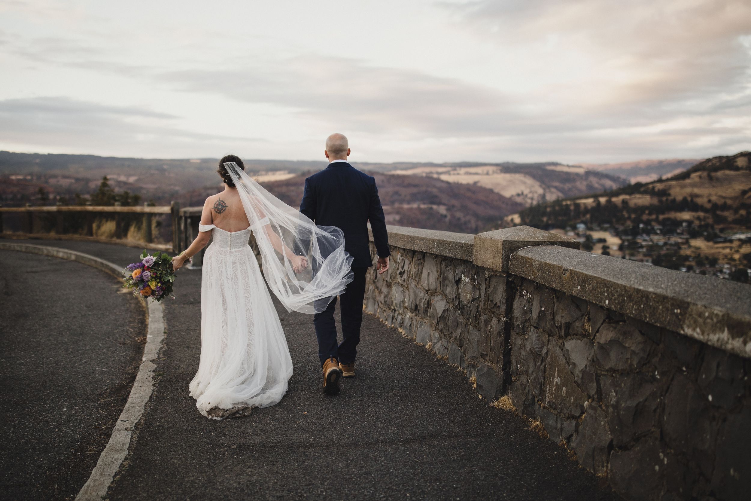 A couple in wedding attire walks along a scenic mountain road with a stone wall and dramatic cloudy sky.