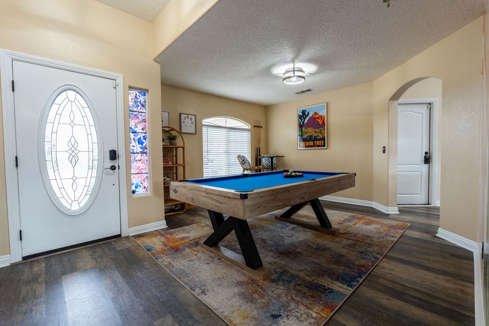 A panoramic view of a game room with a pool table, white walls, and an oval window letting in natural light.