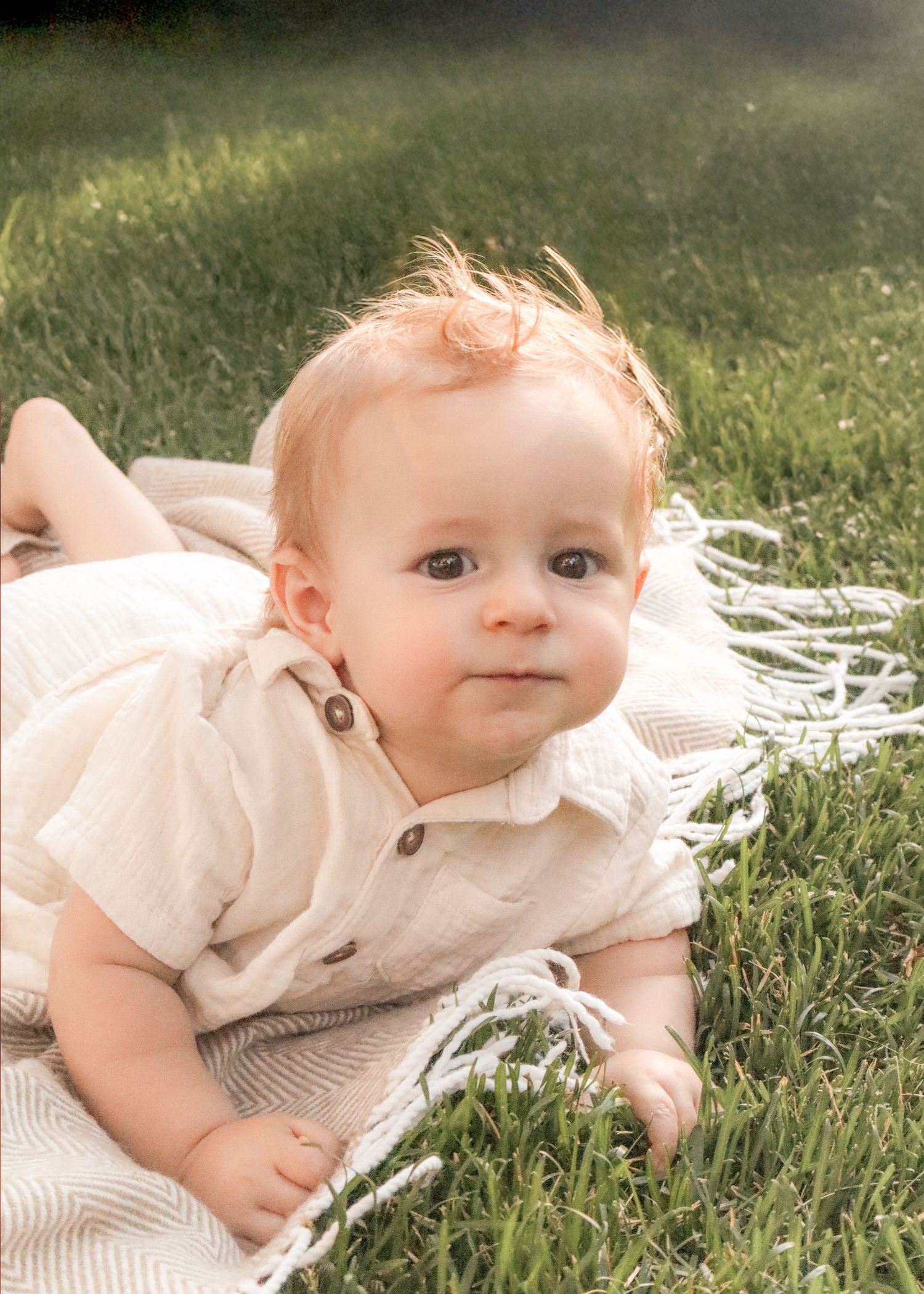 A baby in a cream-colored outfit lies on green grass during a sunny outdoor photo session.
