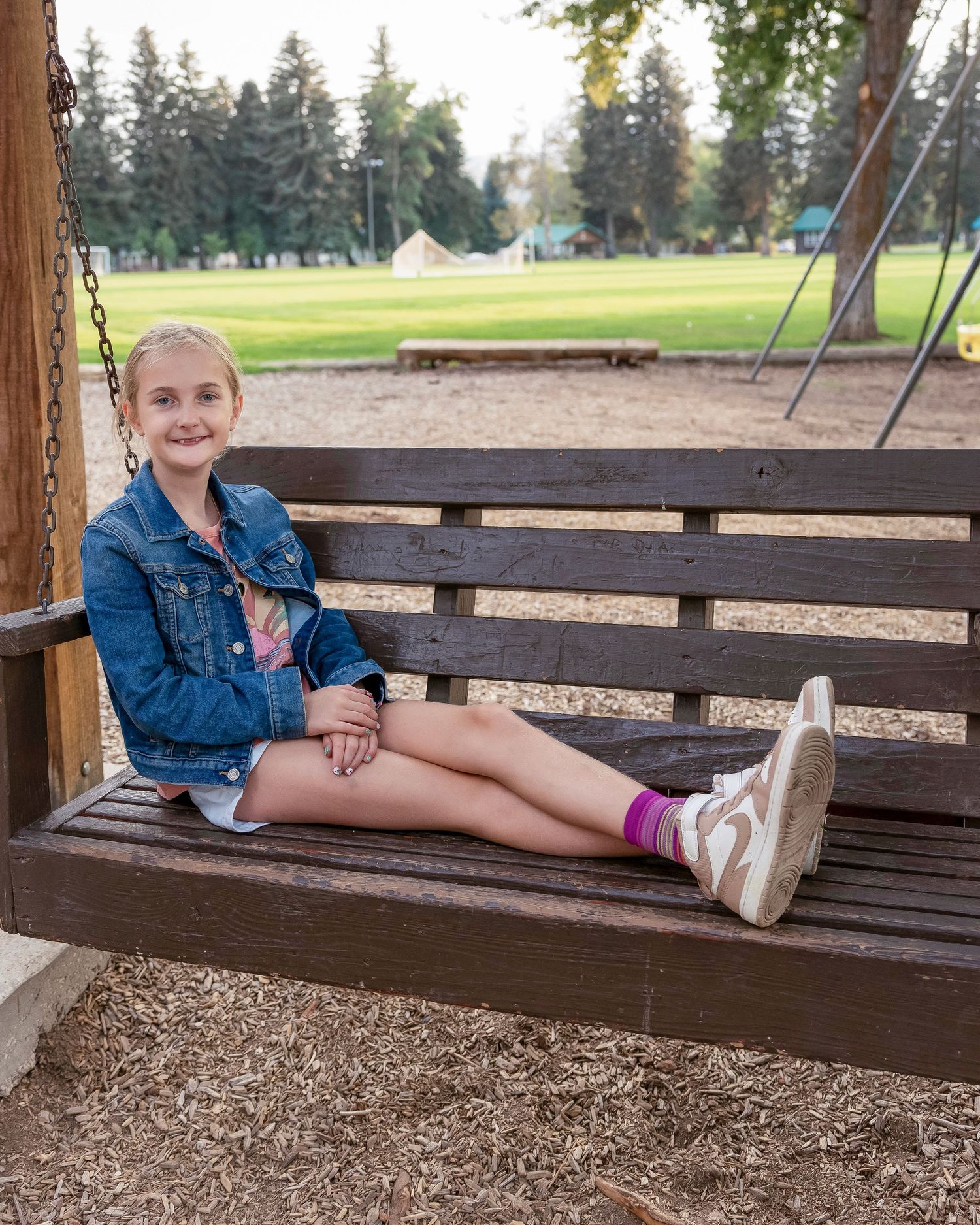 A young person in a denim jacket sits casually on a wooden park swing in an outdoor setting.
