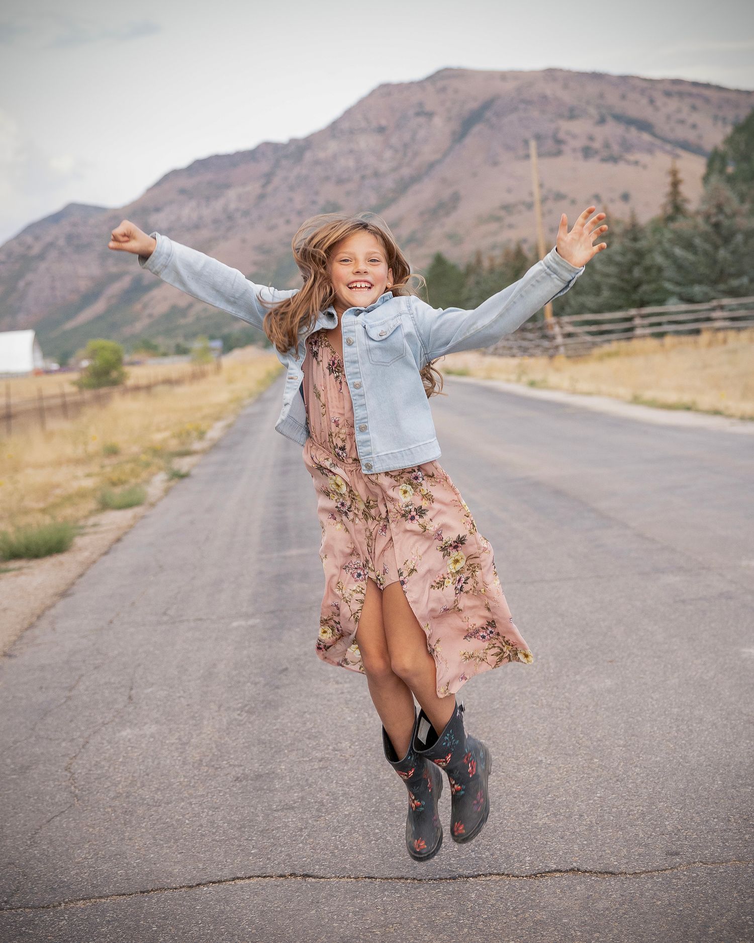 A person in a floral dress and boots dances joyfully on an empty mountain road with scenic hills in the background.