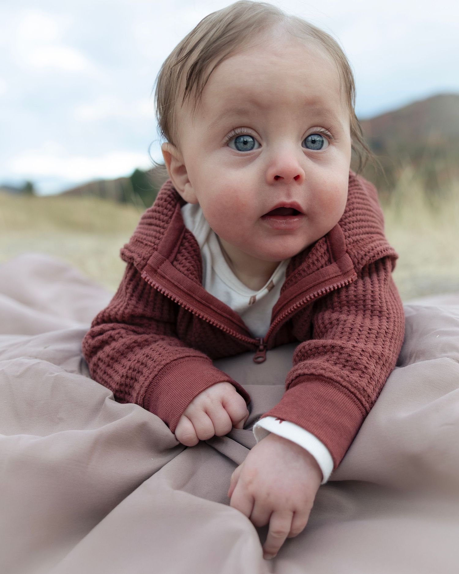 Baby wearing burgundy knit sweater lying on soft fabric with bright blue eyes looking upward.