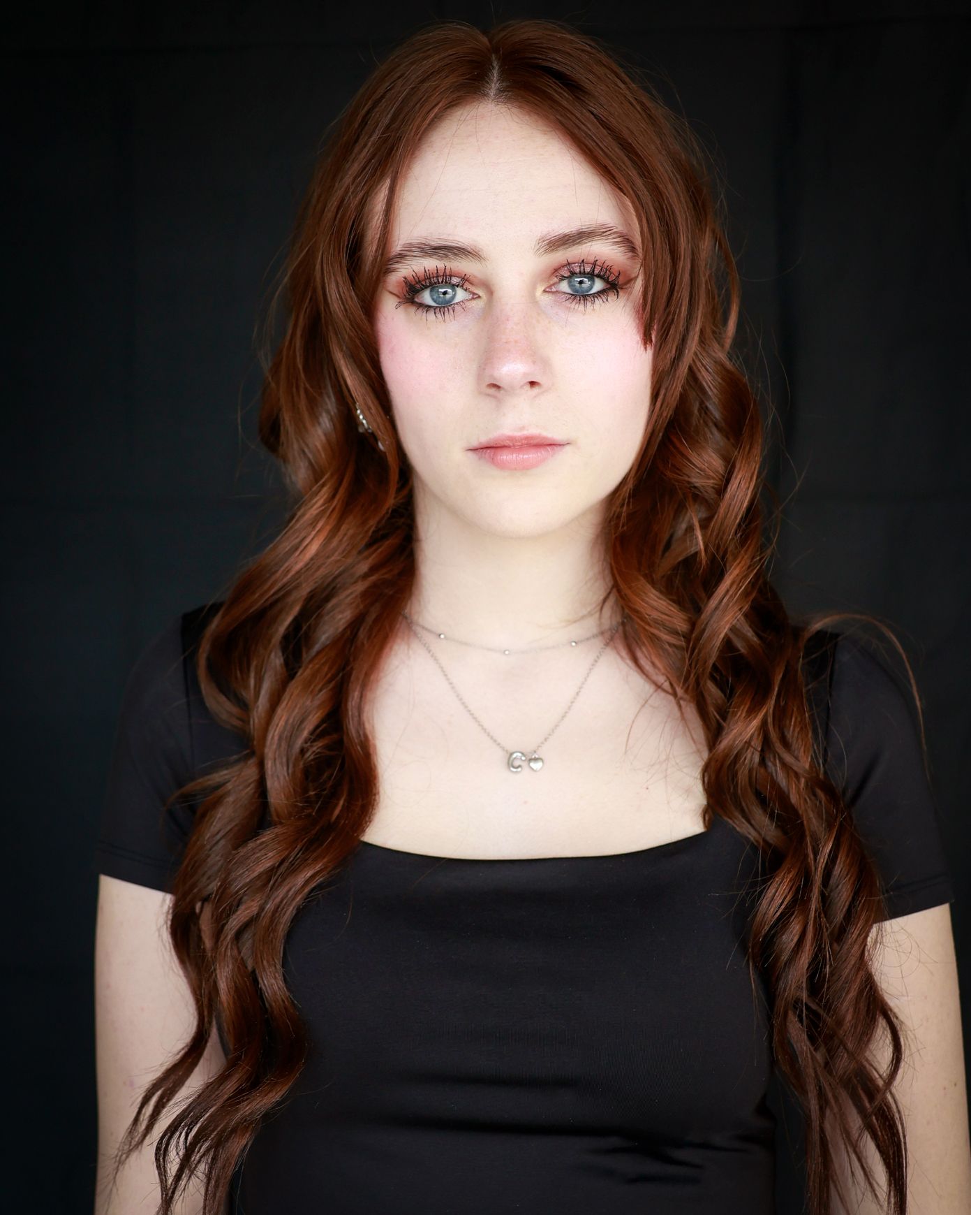 Young woman with long wavy auburn hair and blue eyes wearing a black top and silver necklace against dark background.
