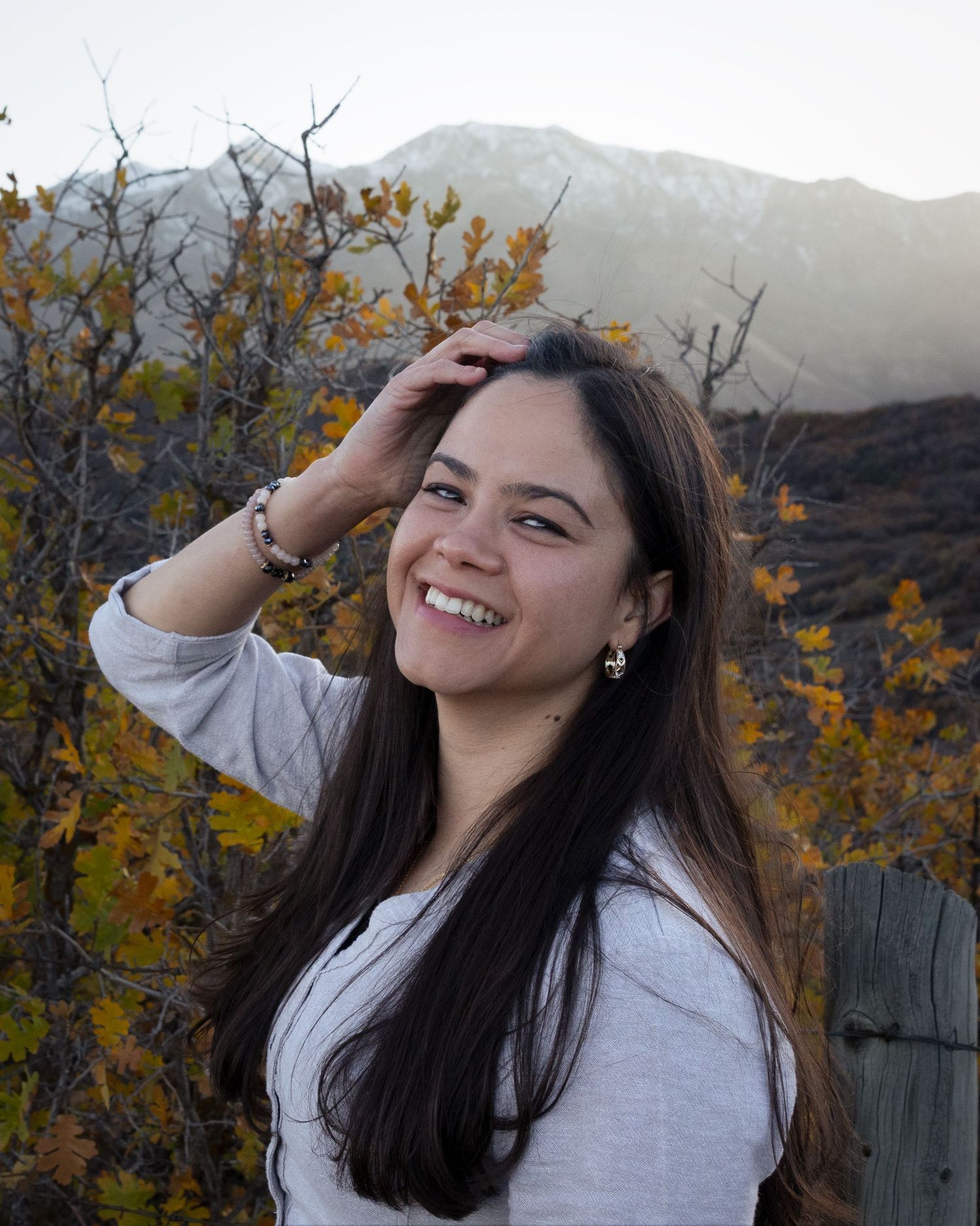 A woman in a white shirt poses joyfully against an autumn mountain backdrop with golden foliage.