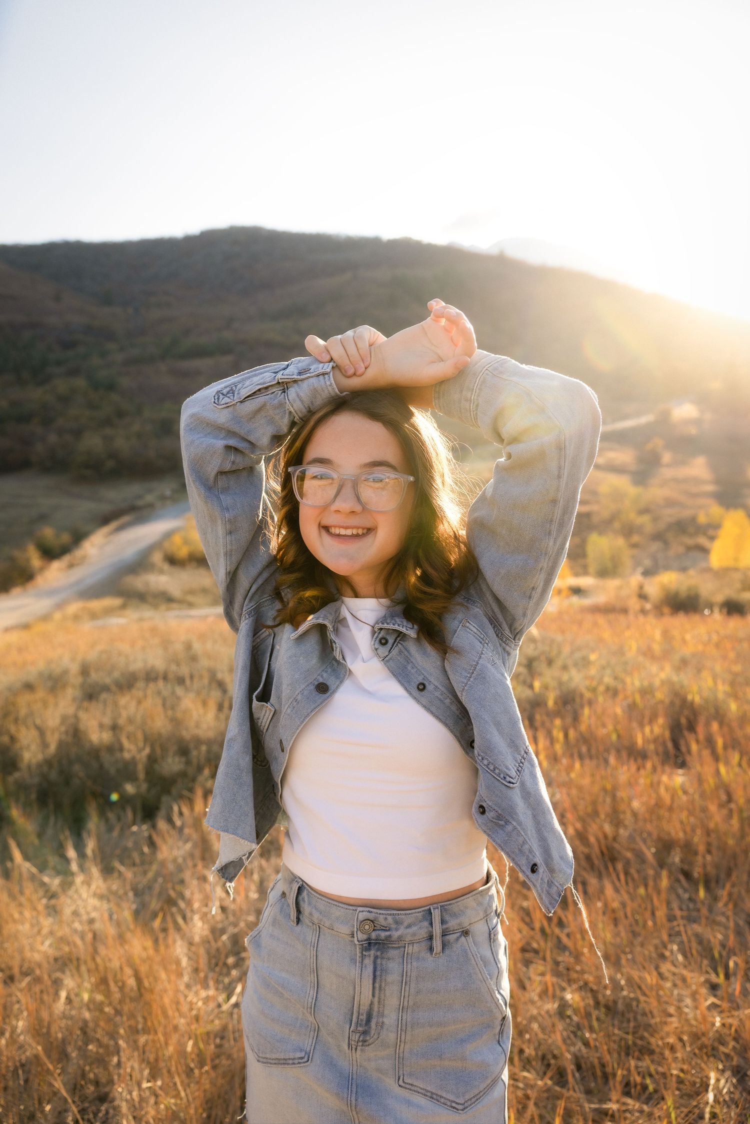 A person in denim overalls poses playfully in a golden field at sunset with mountains in the background.