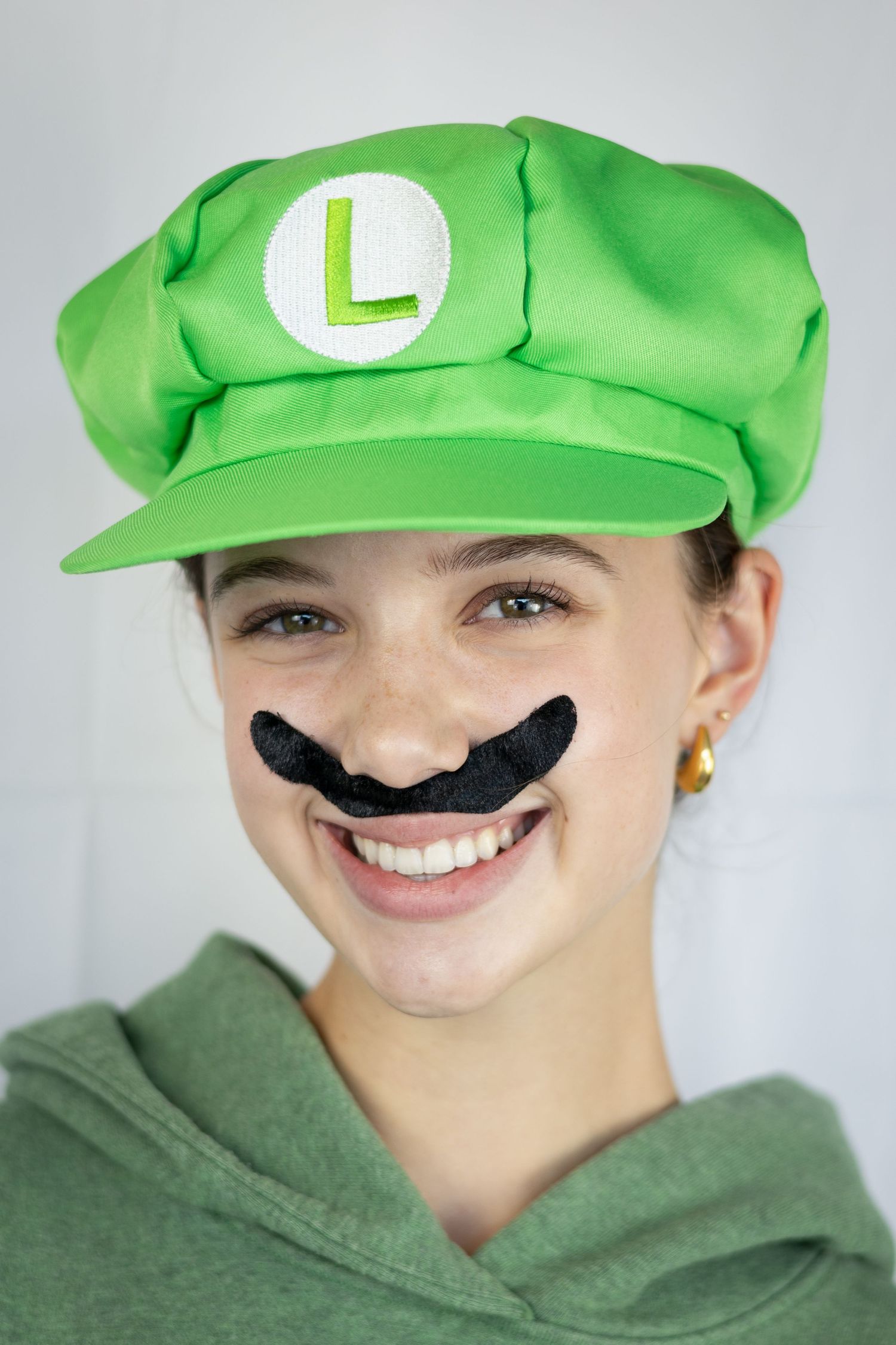 Someone wearing a green Luigi costume cap and fake black mustache smiles while dressed in a green hoodie.