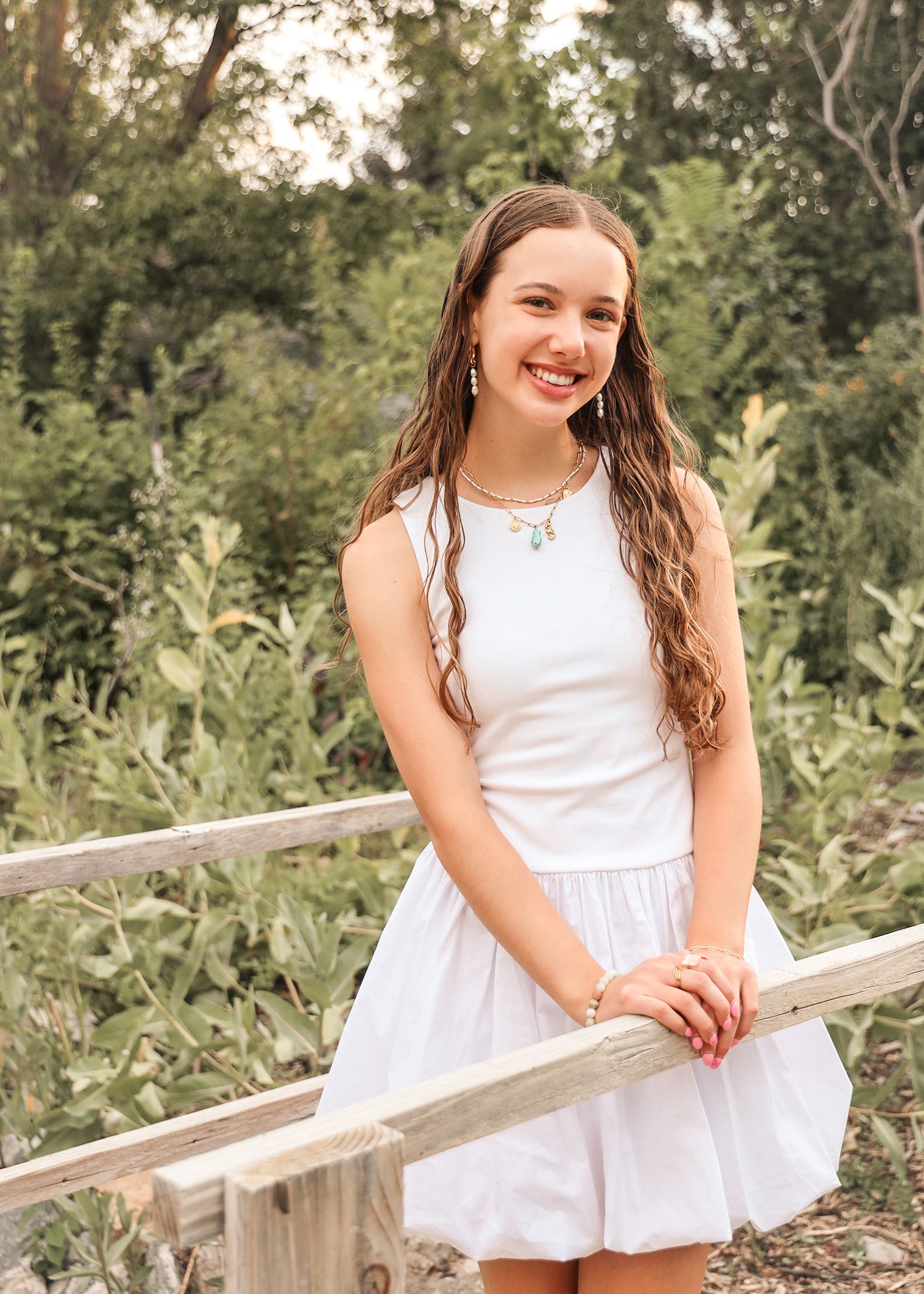 A person in a white summer dress poses outdoors on wooden steps surrounded by lush greenery.