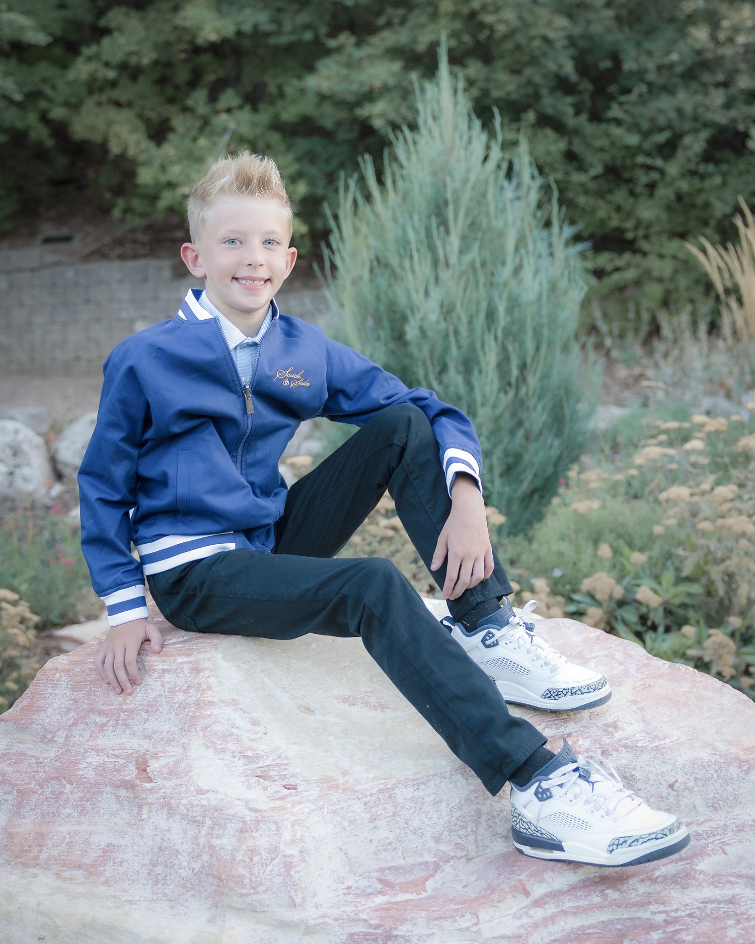 A young person in a blue varsity jacket and jeans sits casually on a large rock with ornamental shrubs in the background.