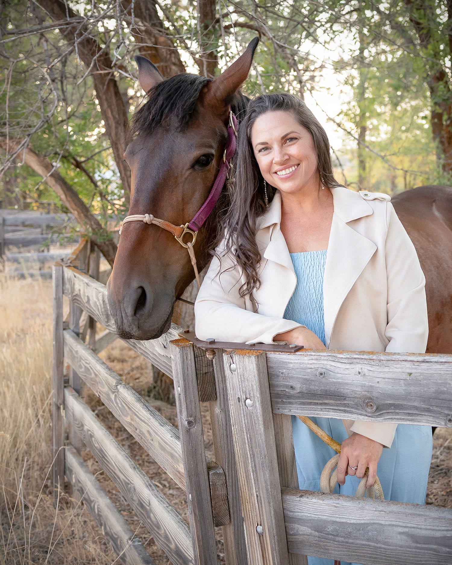 A brown horse and person in a cream coat lean against a wooden fence in an outdoor autumn setting.