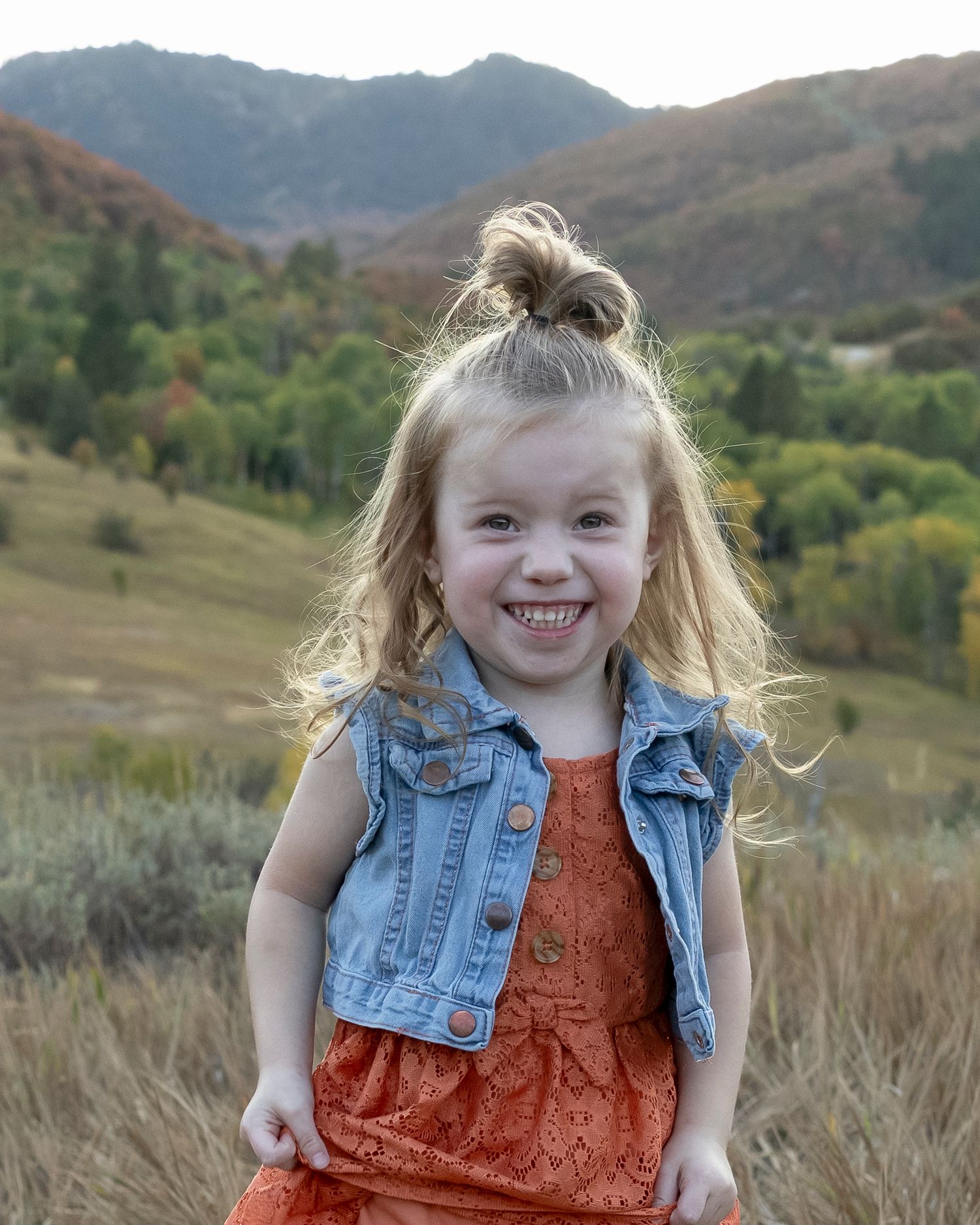 Child in orange dress and denim vest poses outdoors against mountain backdrop in series of candid expressions.