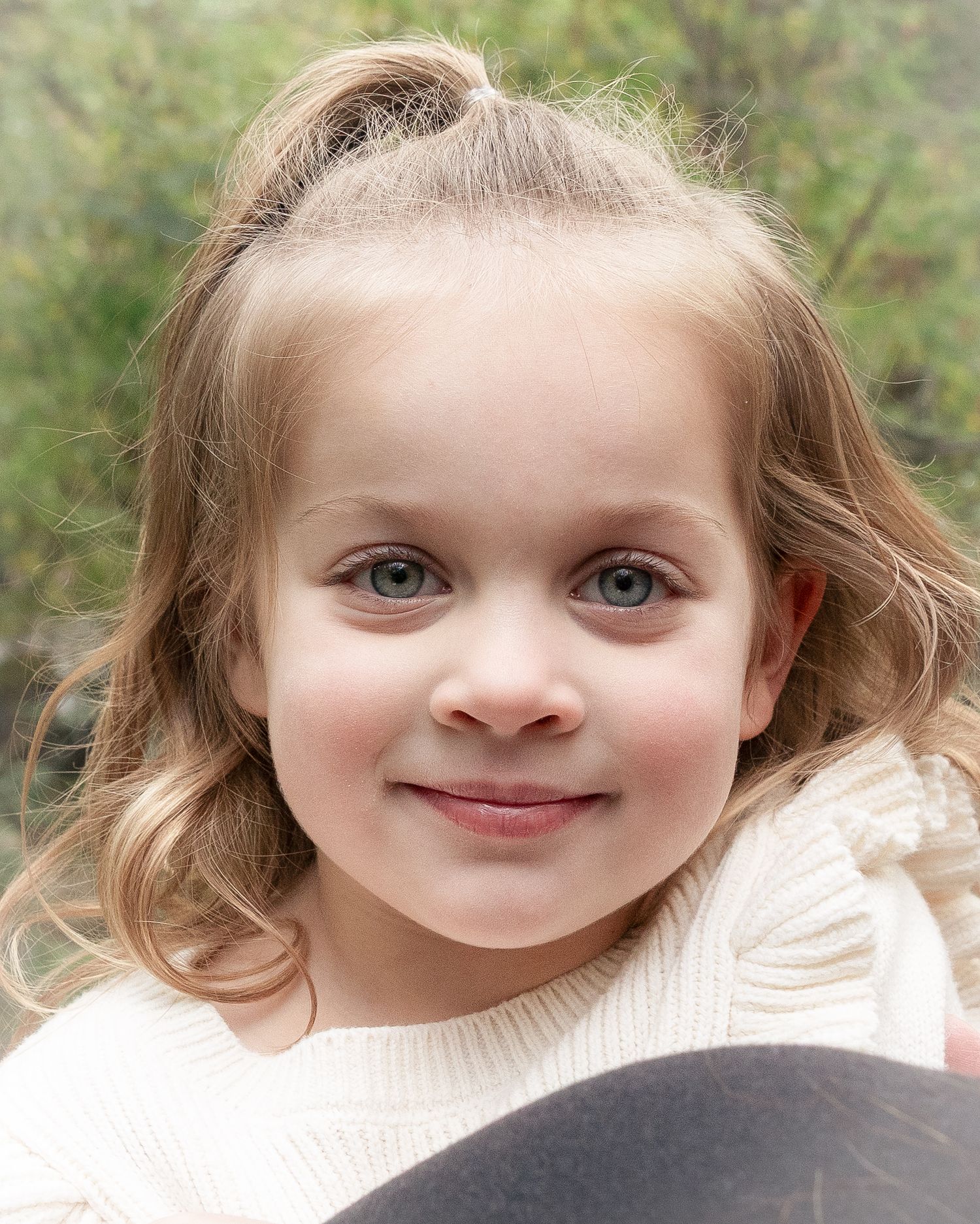 A child with curly blonde hair and bright blue eyes smiles warmly at the camera in a white shirt.