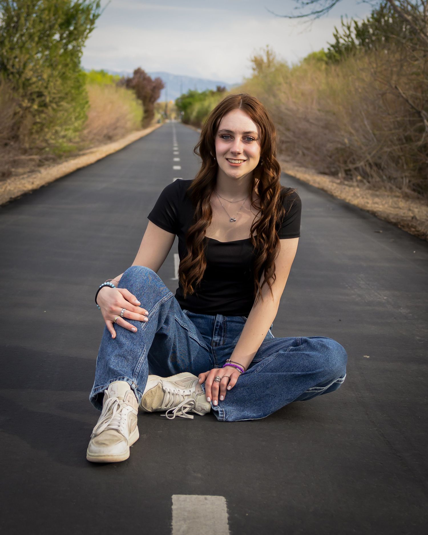 Young woman with long auburn hair sits cross-legged on a quiet road wearing black top and jeans, smiling confidently.