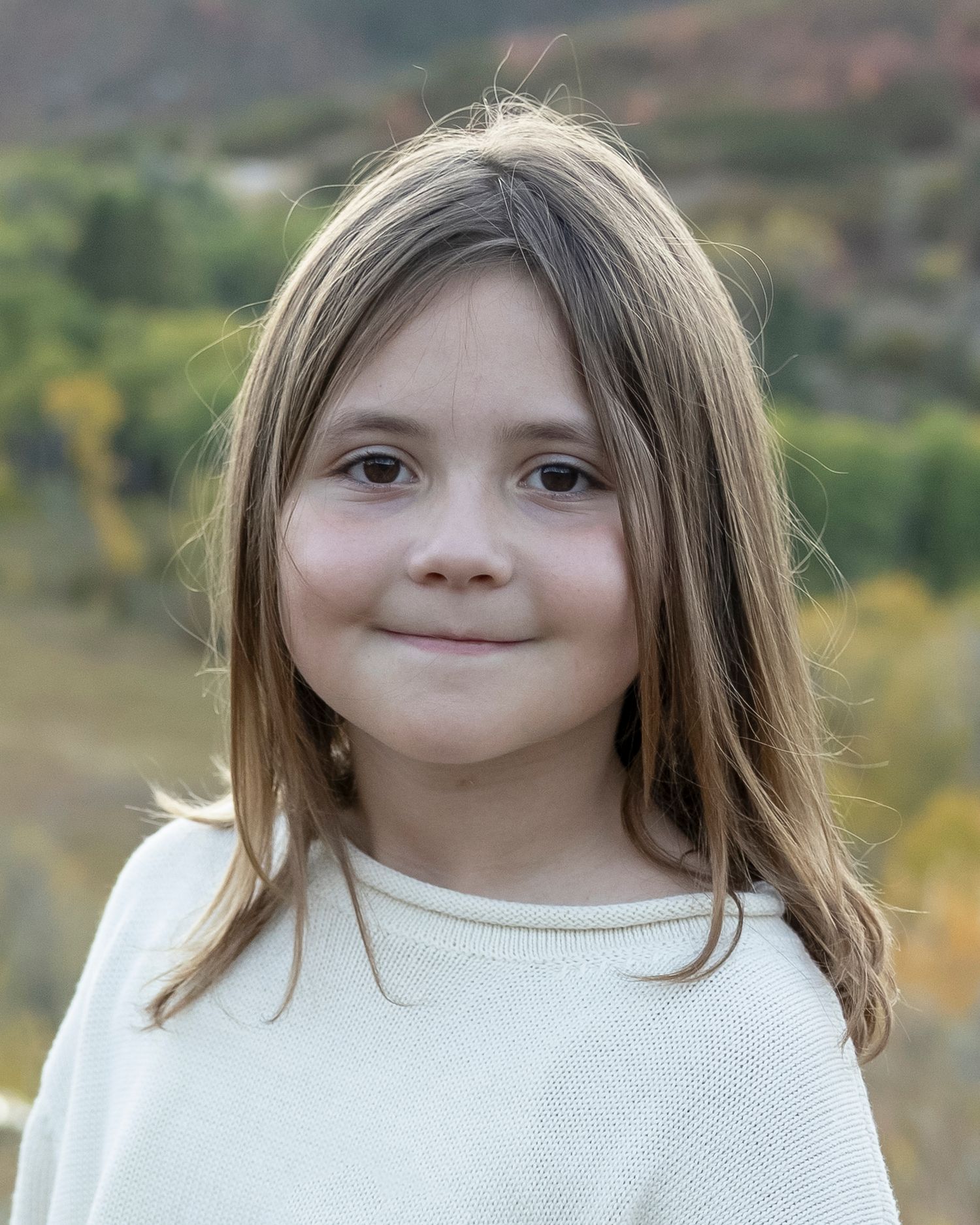 Child wearing a white sweater poses outdoors with autumn foliage in the background.