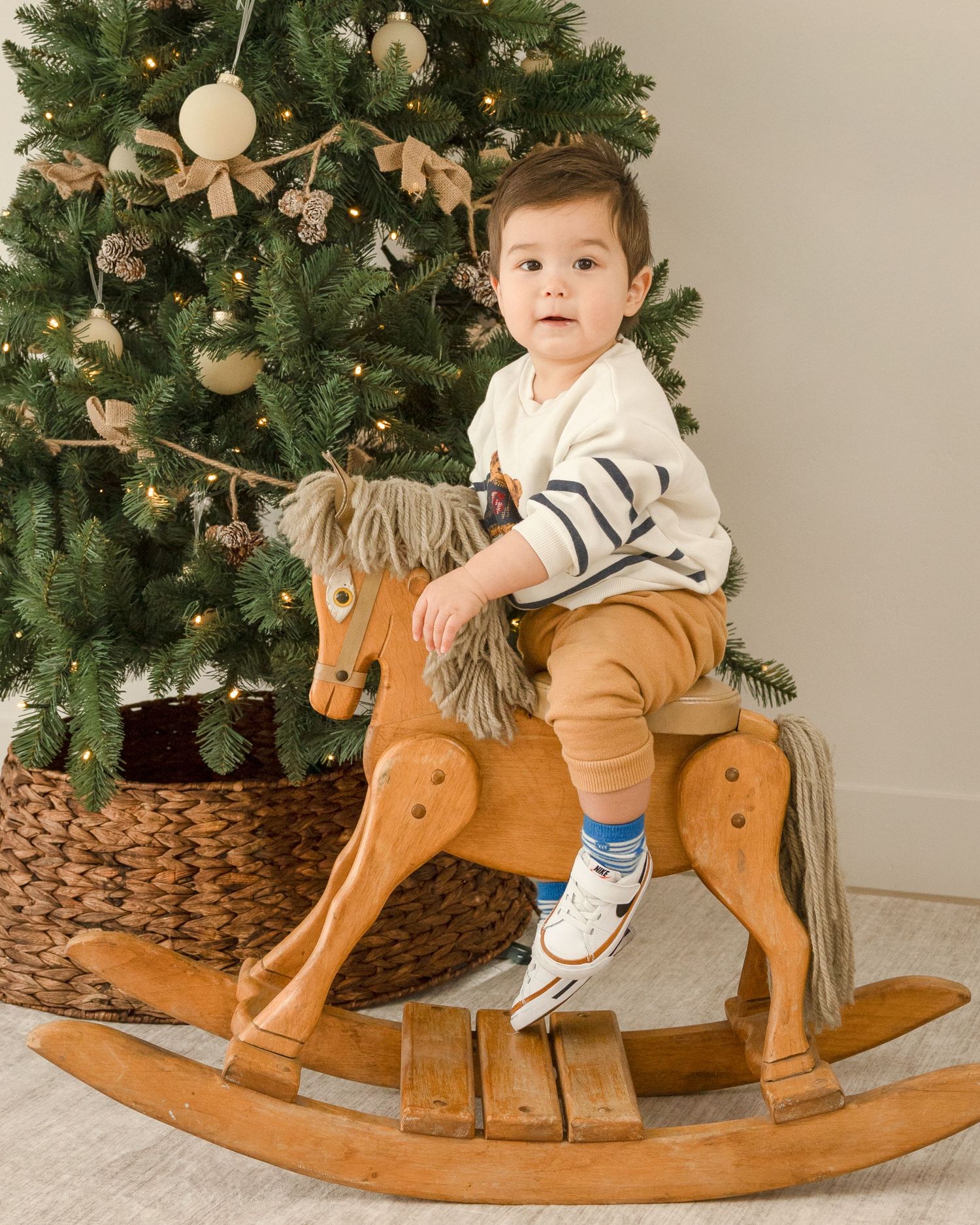 A child sits on a wooden rocking horse in front of a decorated Christmas tree with white and gold ornaments.