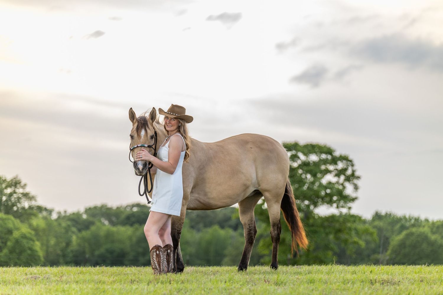 girl standing next to her horse on a hill at sunset wearing white dress and cowboy hat