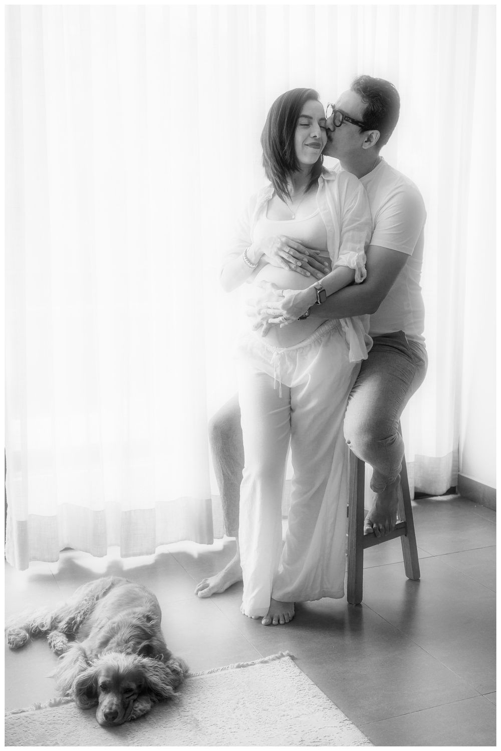Black and white maternity photo of couple embracing near window with dog resting on floor below.