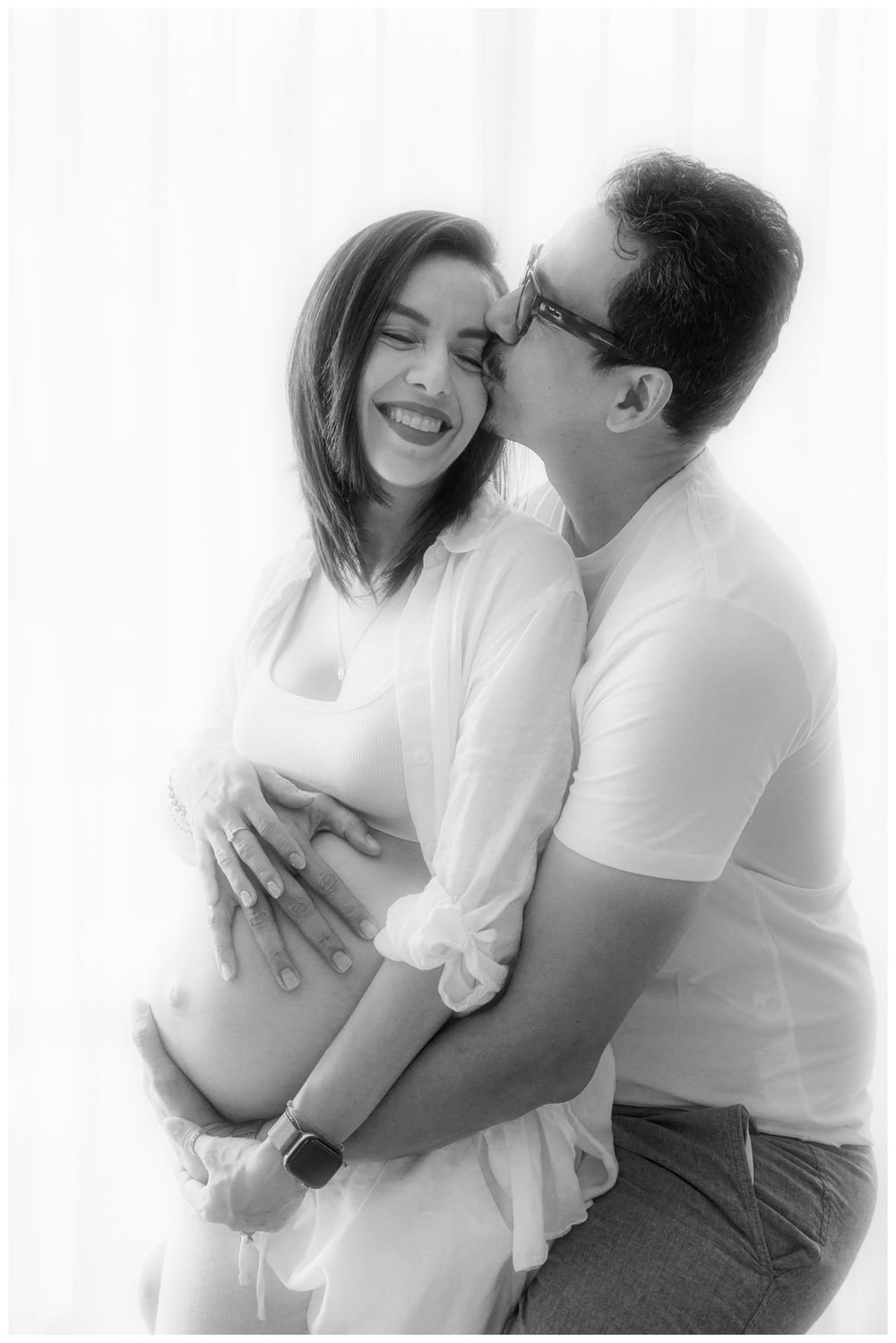 Black and white maternity photo of a smiling pregnant woman being kissed on cheek by her partner.